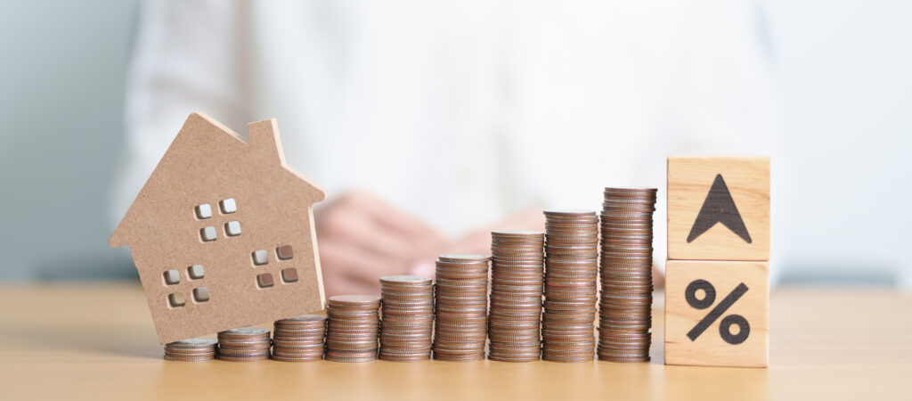 Rostoucí hromádky mincí vedle dřevěného domku a symbolu procenta, znázorňující nárůst daně z nemovitostí.